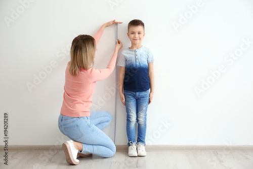Mother measuring height of little boy near light wall