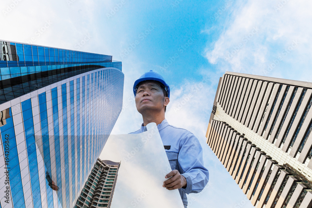 Double exposure engineer wearing blue helmet and holding blueprint looking up with skyscraper background Double exposure engineer wearing blue helmet and holding blueprint looking up with skyscraper background
