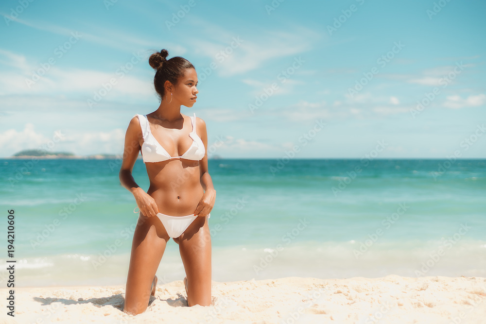 Sexy young Brazilian female on the beach looking aside while standing on knees on sand; with teal sea, horizon, island in blurry background; with copy space place for your logo or advertising message