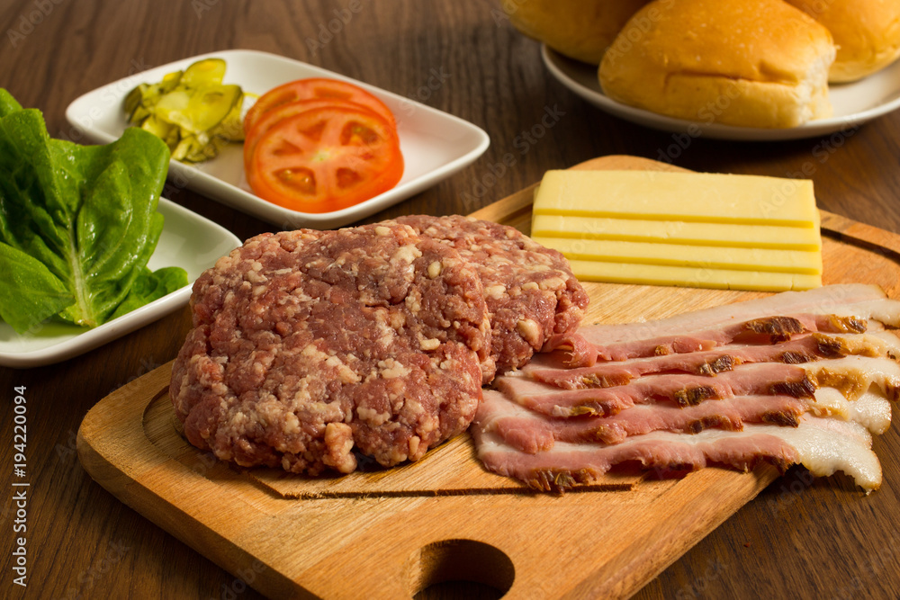 Ingredients for cooking burgers. Raw ground beef meat cutlets on wooden chopping board, tomatoes, greens, pickles, ketchup, cheese,  over wooden background 