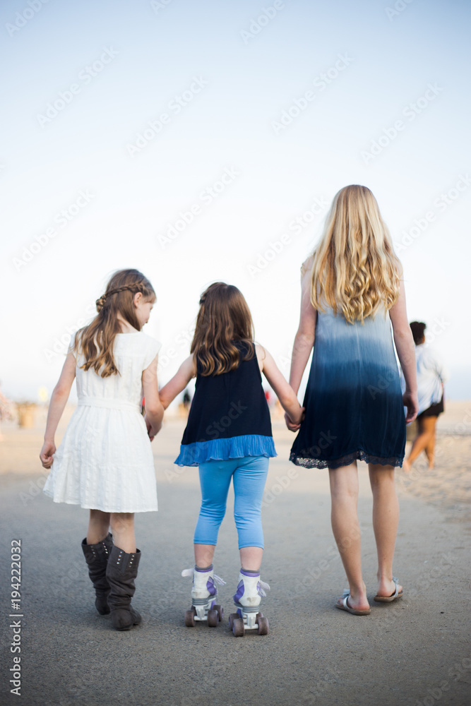 Two older sisters helping younger sister rollerskate on a sidewalk by the beach Two older sisters helping younger sister rollerskate on a sidewalk by the beach