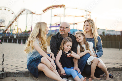Family of five sitting on boardwalk looking at each other with rollercoaster and ferris wheel in the background