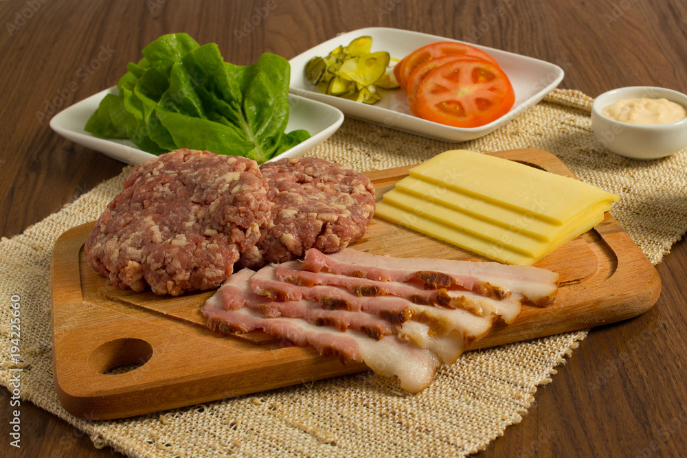 Ingredients for cooking burgers. Raw ground beef meat cutlets on wooden chopping board, tomatoes, greens, pickles, ketchup, cheese, over wooden background Ingredients for cooking burgers. Raw ground beef meat cutlets on wooden chopping board, tomatoes, greens, pickles, ketchup, cheese, over wooden background
