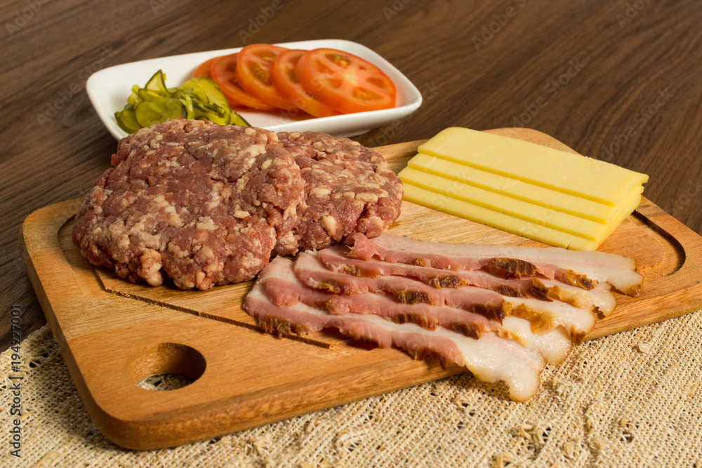 Ingredients for cooking burgers. Raw ground beef meat cutlets on wooden chopping board, tomatoes, greens, pickles, ketchup, cheese, over wooden background Ingredients for cooking burgers. Raw ground beef meat cutlets on wooden chopping board, tomatoes, greens, pickles, ketchup, cheese, over wooden background