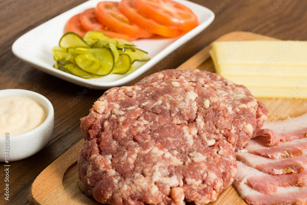 Ingredients for cooking burgers. Raw ground beef meat cutlets on wooden chopping board, tomatoes, greens, pickles, ketchup, cheese, over wooden background - Top View Ingredients for cooking burgers. Raw ground beef meat cutlets on wooden chopping board, tomatoes, greens, pickles, ketchup, cheese, over wooden background - Top View
