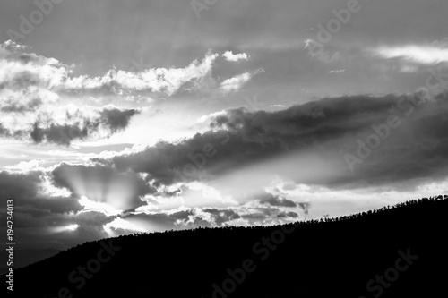 Sunset over a trees line on a hill, with sun rays coming out through some clouds