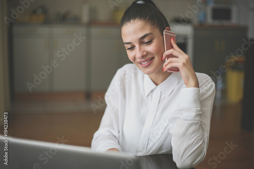 Young smiling business woman working at home. Woman using laptop and talking on smart phone.