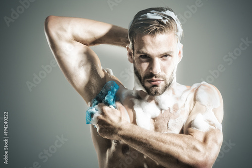 Attractive stylish bearded macho model with fashion hair takes shower with soap sponge. Bodybuilder, sexy strong man washing with foam sponge after workout.