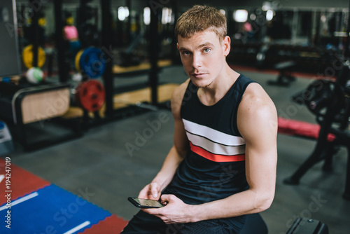 Horizontal shot of athletic man in the gym using smart phone for sport application doing fitness exercise. Sporty male sitting on a brench in the fitness club with copy space for text or advertising.