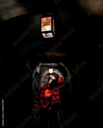 Model in red fur jacket poses under the lantern in the dark tunnel