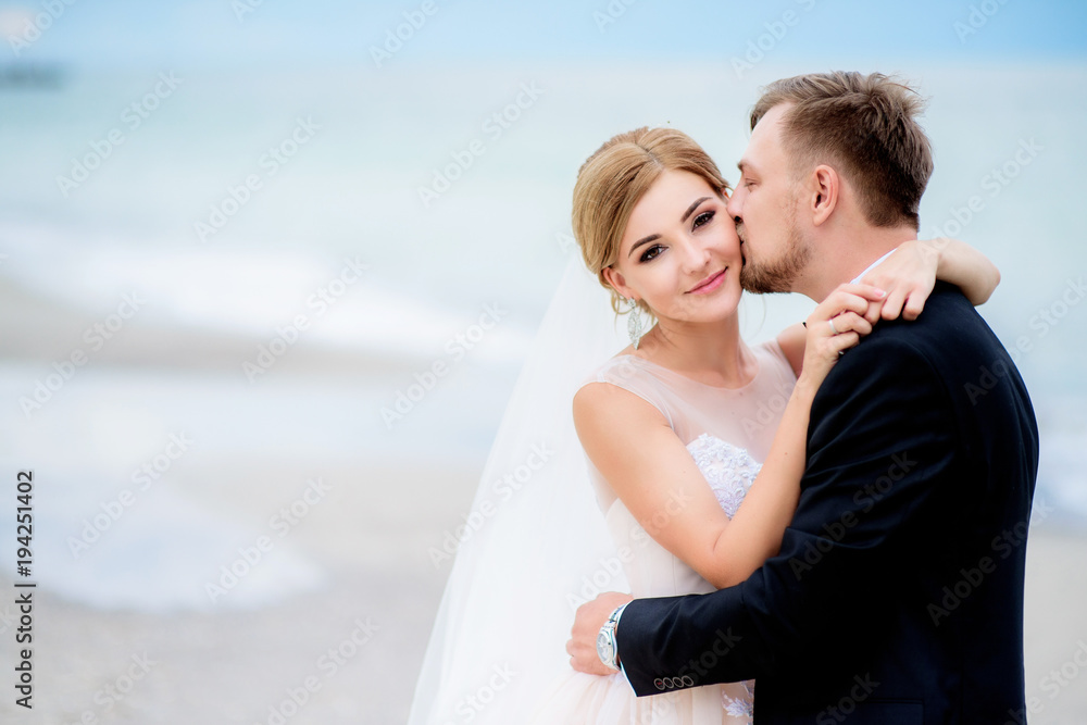 Dreamy wedding couple hug each other tender standing on the beach with sea waves splashing behind them Dreamy wedding couple hug each other tender standing on the beach with sea waves splashing behind them