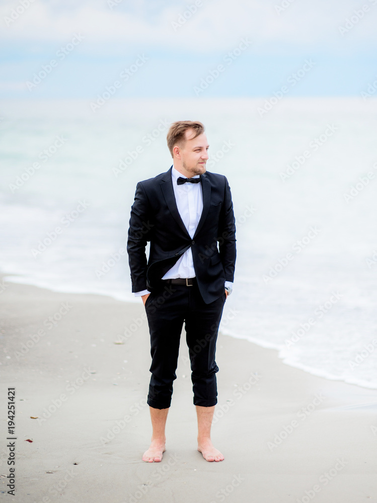Wind shakes groom's hair standing on the beach with waves splashing behind him Wind shakes groom's hair standing on the beach with waves splashing behind him