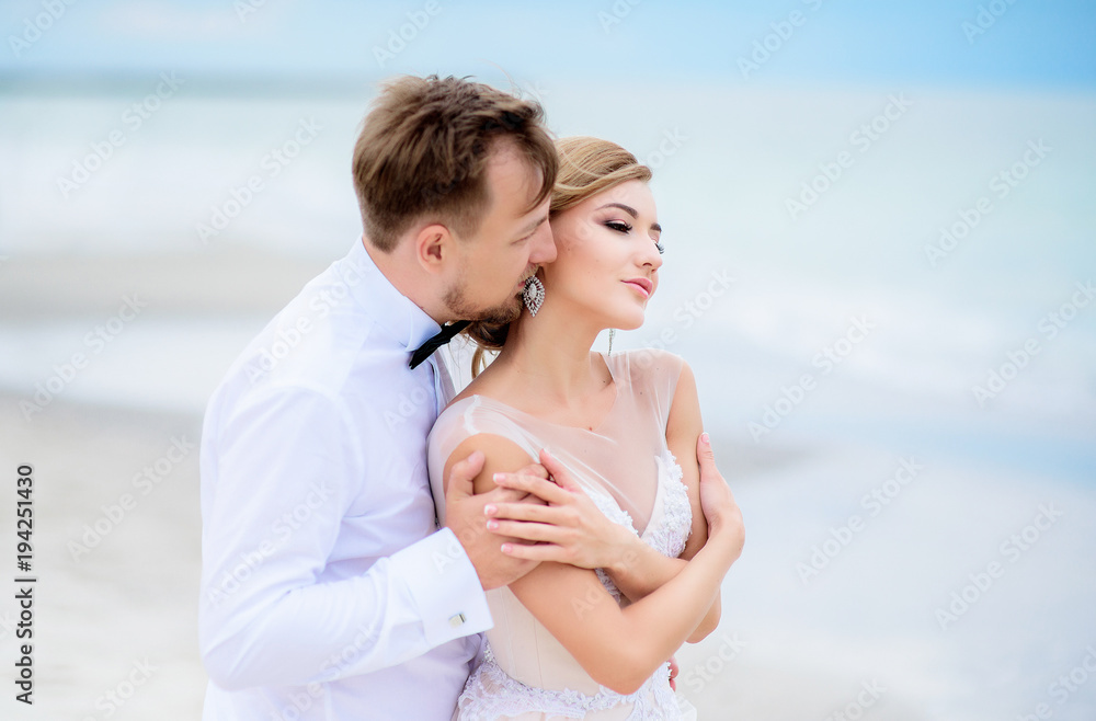 Dreamy wedding couple hug each other tender standing on the beach with sea waves splashing behind them