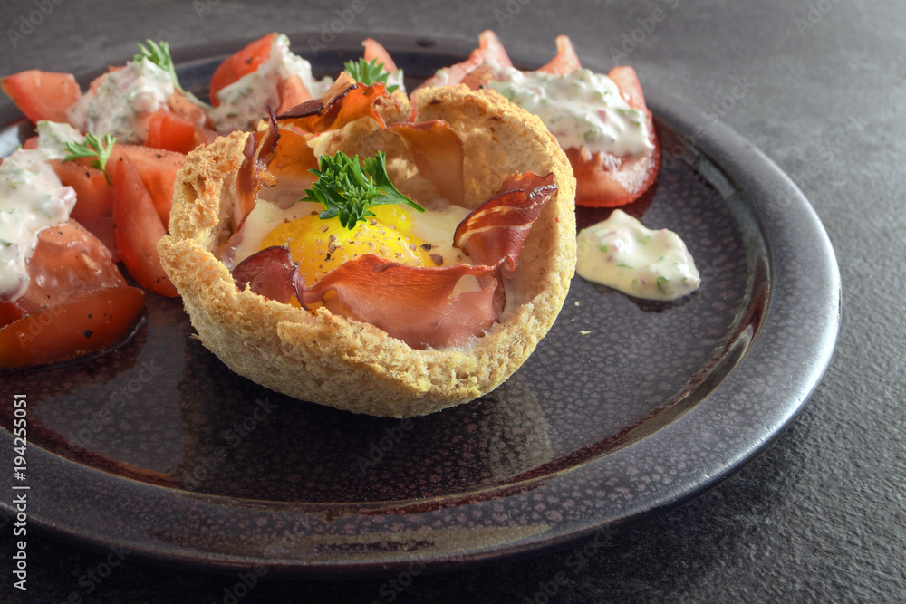 muffin from egg and bacon in toasted bread, tomatoes and cheese cream with herbs on a dark plate on rustic slate, creative breakfast, brunch or snack