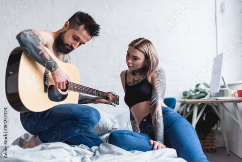 tattooed boyfriend playing guitar for girlfriend in bedroom