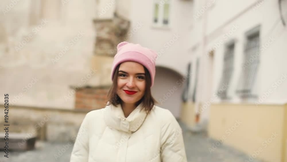 Brunette pretty young women portrait in cute pink hat and red lips turns around, smiles, flirts and looks in to the camera. Old city background. Cold season. Slow motion. Brunette pretty young women portrait in cute pink hat and red lips turns around, smiles, flirts and looks in to the camera. Old city background. Cold season. Slow motion.
