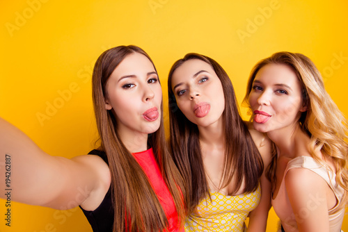 Self portrait of charming, funky, nice, attractive, pretty, crazy girls making selfie on smart phone over yellow background, showing tongue out to the front camera, birthday party, women's day
