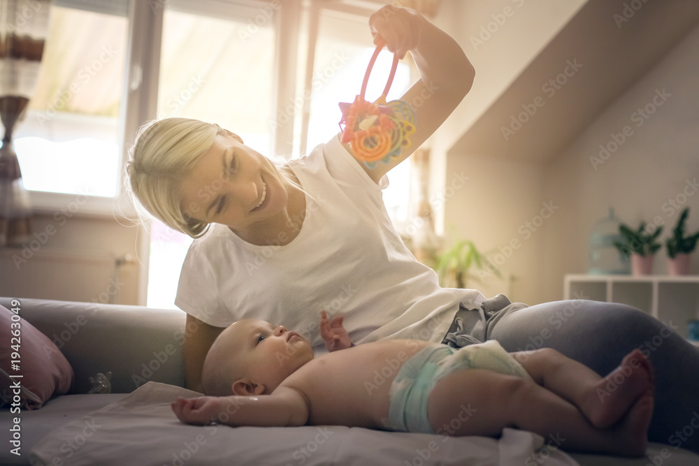 Cheerful mother plying with her little baby boy at home and holding toys.