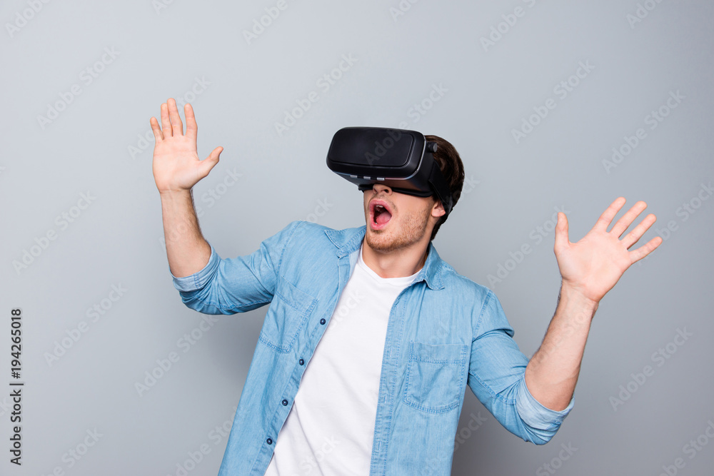 Close up of impressed, shocked, handsome, attractive guy in jeans shirt wearing virtual reality goggles, getting experience using VR-headset, gesticulating, gesturing with hands over gray background Close up of impressed, shocked, handsome, attractive guy in jeans shirt wearing virtual reality goggles, getting experience using VR-headset, gesticulating, gesturing with hands over gray background