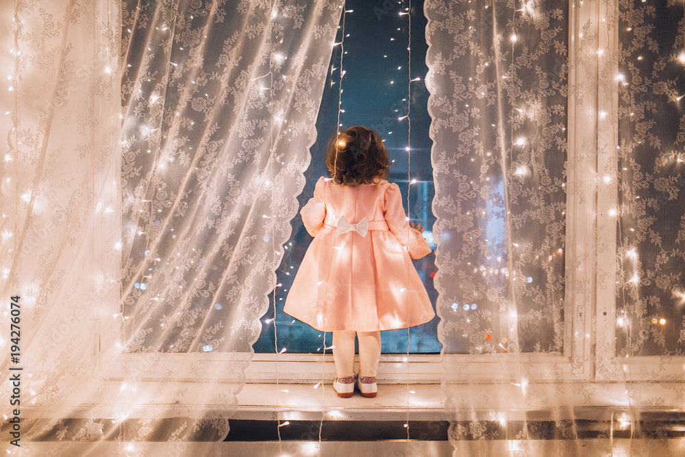 Cute child girl stay near the window and looking at the stars. Little kid stay at windowsill around garlands. Child like in fairytale. Curly child enchanted stars and sky.