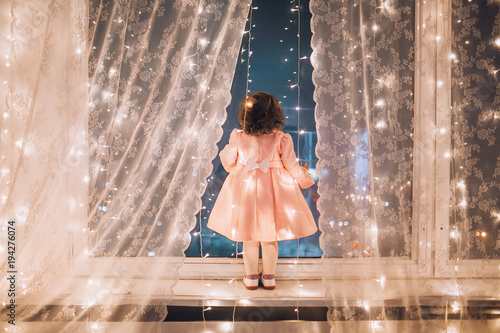 Obraz na płótnie Cute child girl stay near the window and looking at the stars. Little kid stay at windowsill around garlands. Child like in fairytale. Curly child enchanted stars and sky. w salonie