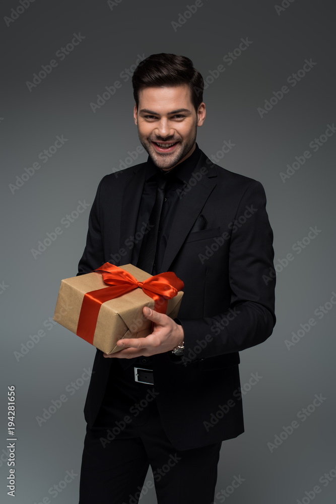 Young smiling male in formal suit holding gift box isolated on grey, international womens day concept Young smiling male in formal suit holding gift box isolated on grey, international womens day concept