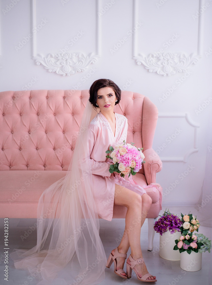 Beautiful thoughtful bride sits in pink silk robe and veil on the luxury pink couch with a wedding bouquet Beautiful thoughtful bride sits in pink silk robe and veil on the luxury pink couch with a wedding bouquet