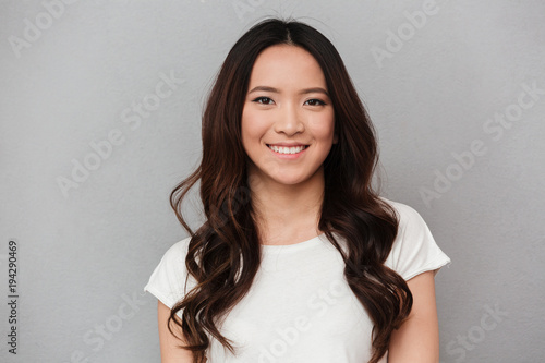 Portrait of asian lovely woman with dark curly hair posing with kind smile, isolated over gray background