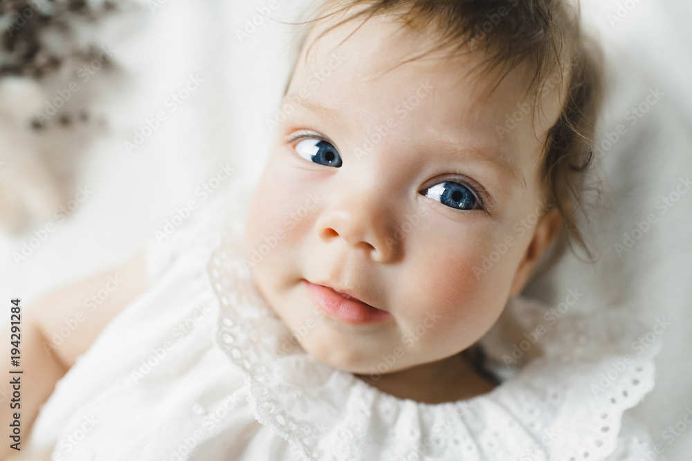 Gorgeous child with blue eyes in white dress lies on white bed in a bright room Gorgeous child with blue eyes in white dress lies on white bed in a bright room
