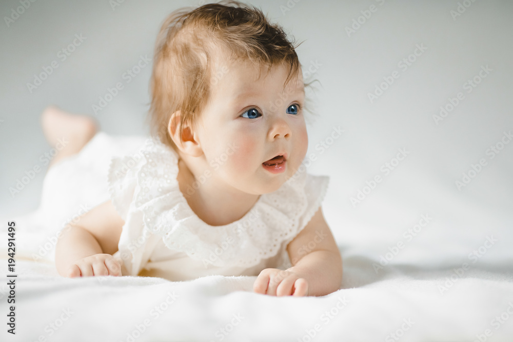 Gorgeous child with blue eyes in white dress lies on white bed in a bright room Gorgeous child with blue eyes in white dress lies on white bed in a bright room