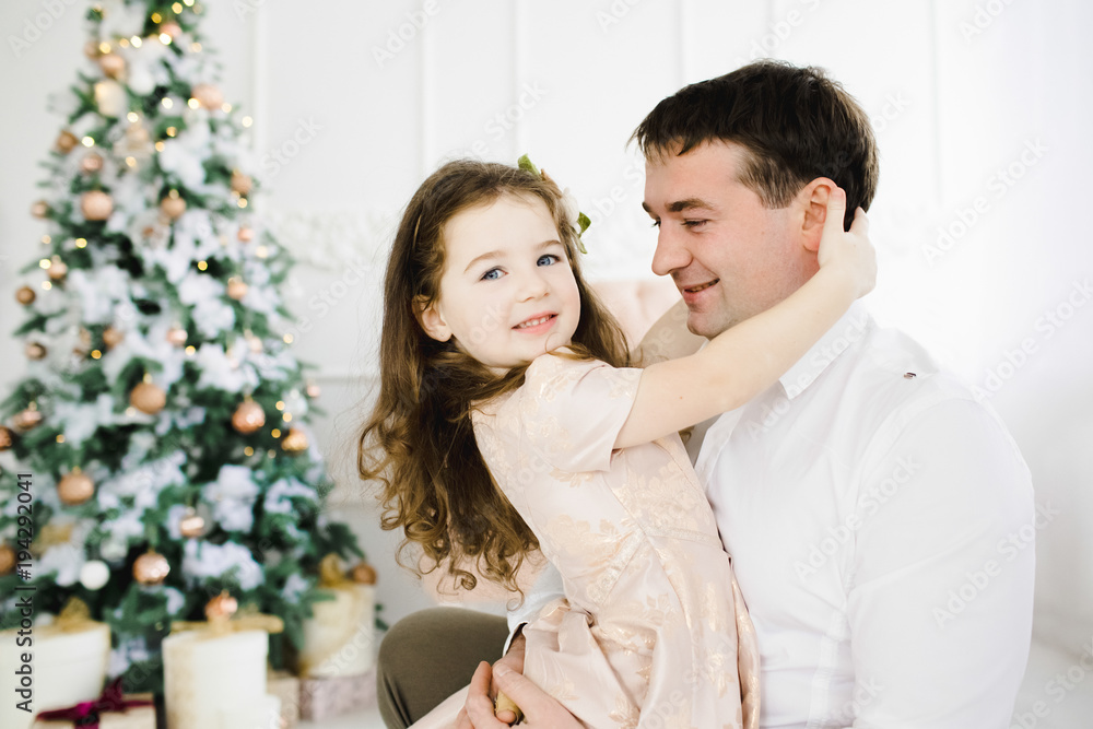 Happy father holds his little pretty girl in peach dress standing before a Christmas tree