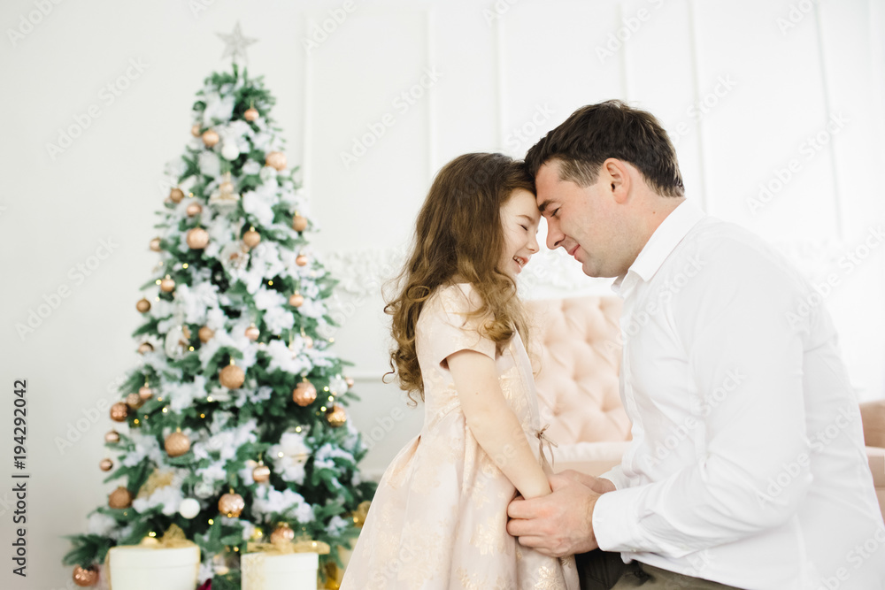 Happy father holds his little pretty girl in peach dress standing before a Christmas tree Happy father holds his little pretty girl in peach dress standing before a Christmas tree