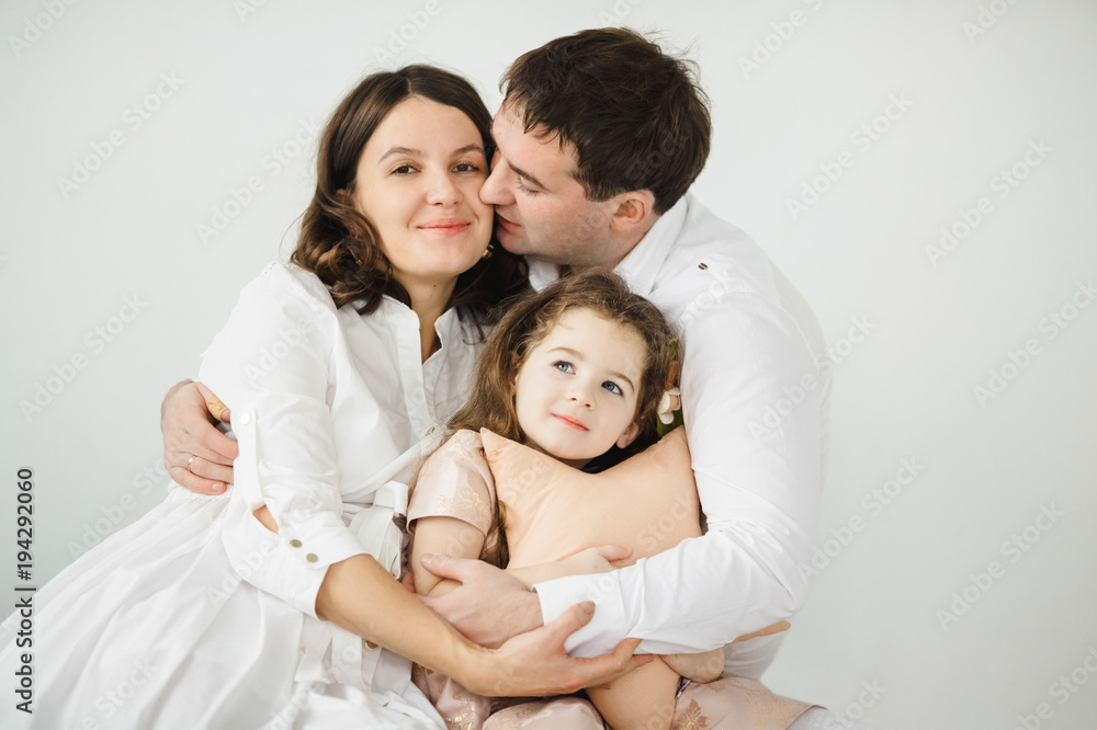 Pregnant woman, her man and their cheerful little daughter in peach dress hug each other tender sitting before a white wall