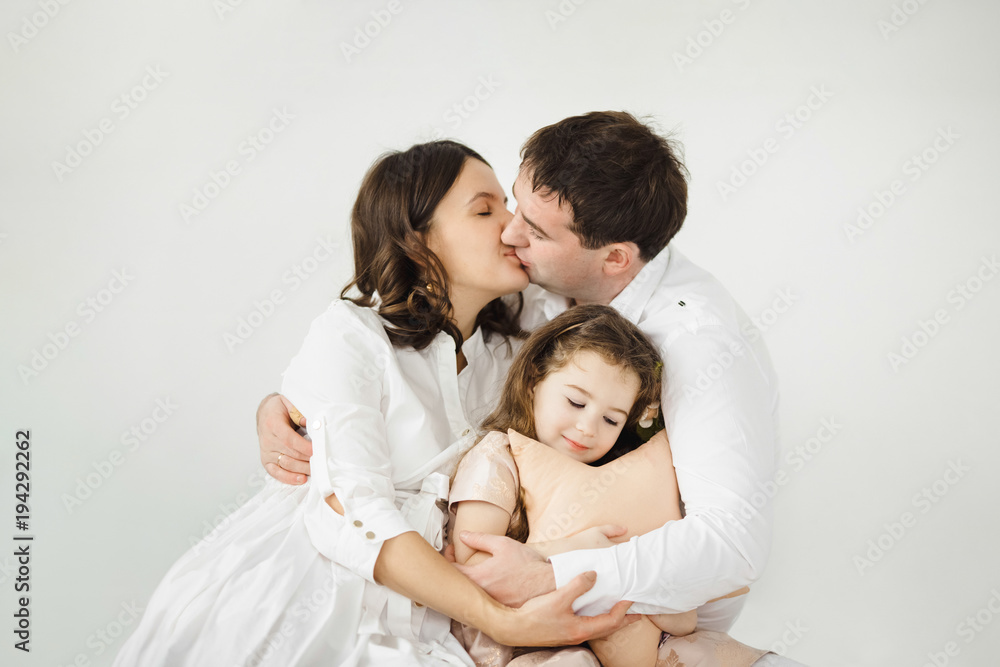 Pregnant woman, her man and their cheerful little daughter in peach dress hug each other tender sitting before a white wall Pregnant woman, her man and their cheerful little daughter in peach dress hug each other tender sitting before a white wall
