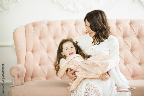 Cheerful mom in a white dress sits with little daughter on a nice peach sofa in luxury white room