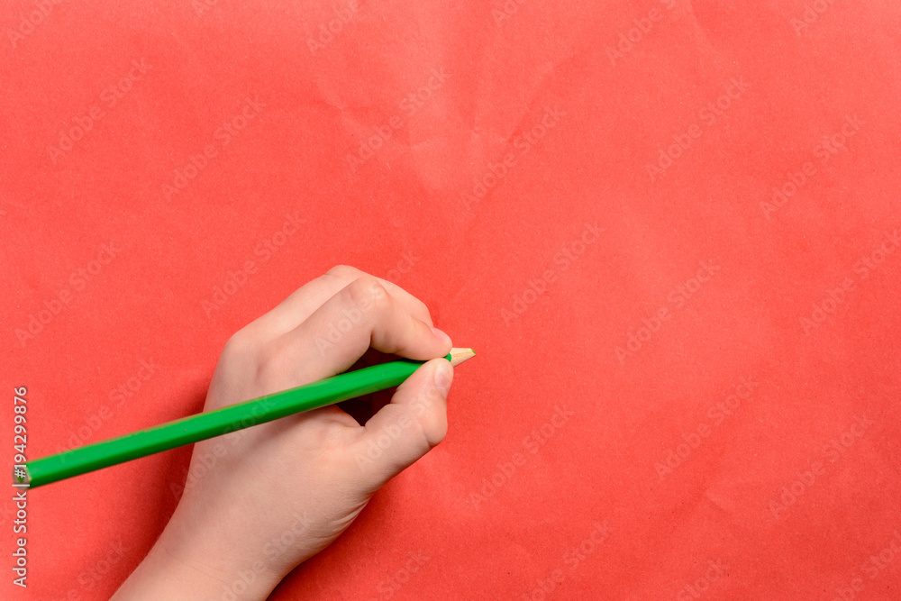 The hand of a young boy holds a green pencil on a red background The hand of a young boy holds a green pencil on a red background