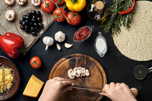 Female hand cut mushrooms on wooden board on kitchen table, around lie ingredients for pizza: vegetables, cheese and spices.