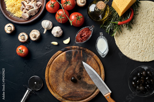 Cutting wooden board with traditional pizza preparation ingredients: cheese, tomatoes sauce, olives, olive oil, pepper, spices. Black texture table background