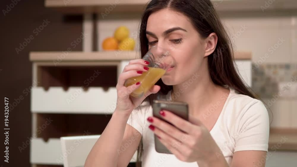 Happy charming brunette texting on the phone, having juice as using the phone, indoor shot in cozy kitchen Happy charming brunette texting on the phone, having juice as using the phone, indoor shot in cozy kitchen
