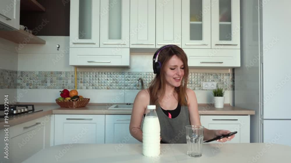 Long-haired woman pouring milk into the glass, singing happily while listening to music on the phone, indoor shot during morning time in kitchen Long-haired woman pouring milk into the glass, singing happily while listening to music on the phone, indoor shot during morning time in kitchen