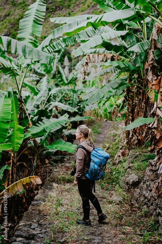 Blond yong women with camera and blue backpack walking through banana plantation on the trekking route to paul valley. Santo antao island. Cape verde photo