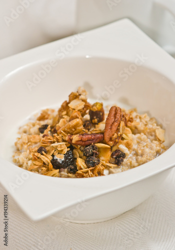 Close up, top view of a square, deep, white bowl of granola with raisins and walnut halves with skim milk on a white place mat, paleo diet