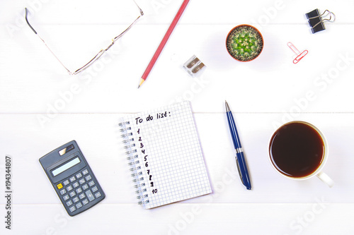 Notepad with text: To do list. White table with calculator, cactus, note paper, coffee mug, pen, glasses.