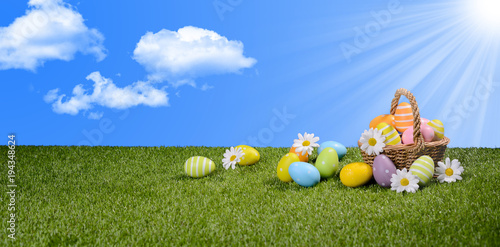 basket of multi colored painted easter eggs on green grass with springtime daisy flowers and blue sky