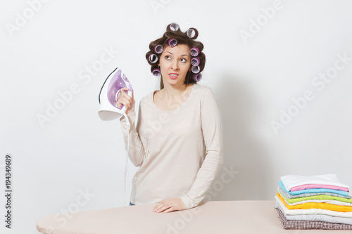 Crazy fun housewife with curlers on hair in light clothes ironing family clothing on ironing board with iron. Woman isolated on white background. Housekeeping concept. Copy space for advertisement.