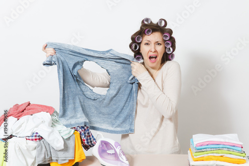 Shocked fun crazy housewife, curlers on hair in light clothes holding burned shirt with hole made by iron, standing at ironing board. Woman isolated on white background. Copy space for advertisement.