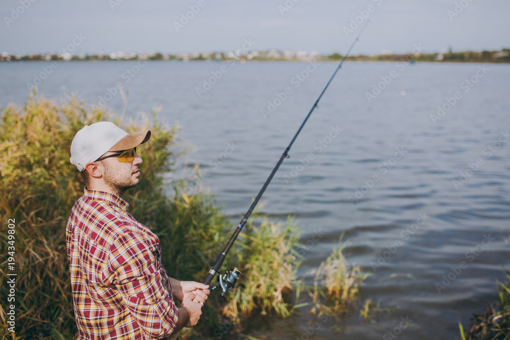 Side view Young unshaven man with a fishing rod in checkered shirt, cap and sunglasses looks into distance on lake from shore near shrubs and reeds. Lifestyle, recreation, fisherman leisure concept. Side view Young unshaven man with a fishing rod in checkered shirt, cap and sunglasses looks into distance on lake from shore near shrubs and reeds. Lifestyle, recreation, fisherman leisure concept.