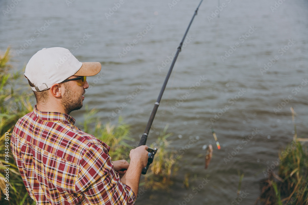 Side view Young unshaven man in checkered shirt, cap and sunglasses pulls out fishing pole with caught fish on lake from shore near shrubs and reeds. Lifestyle, recreation, fisherman leisure concept Side view Young unshaven man in checkered shirt, cap and sunglasses pulls out fishing pole with caught fish on lake from shore near shrubs and reeds. Lifestyle, recreation, fisherman leisure concept