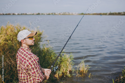 Side view Young unshaven man with a fishing rod in checkered shirt, cap and sunglasses looks into distance on lake from shore near shrubs and reeds. Lifestyle, recreation, fisherman leisure concept.