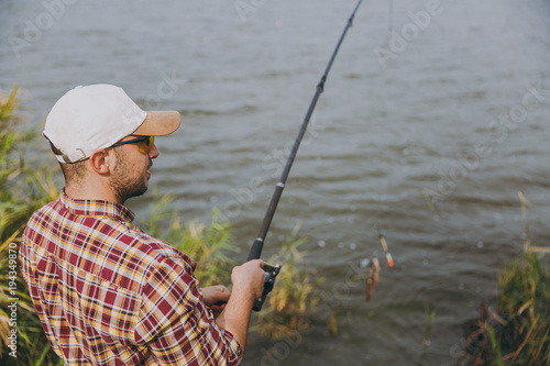 Side view Young unshaven man in checkered shirt, cap and sunglasses pulls out fishing pole with caught fish on lake from shore near shrubs and reeds. Lifestyle, recreation, fisherman leisure concept